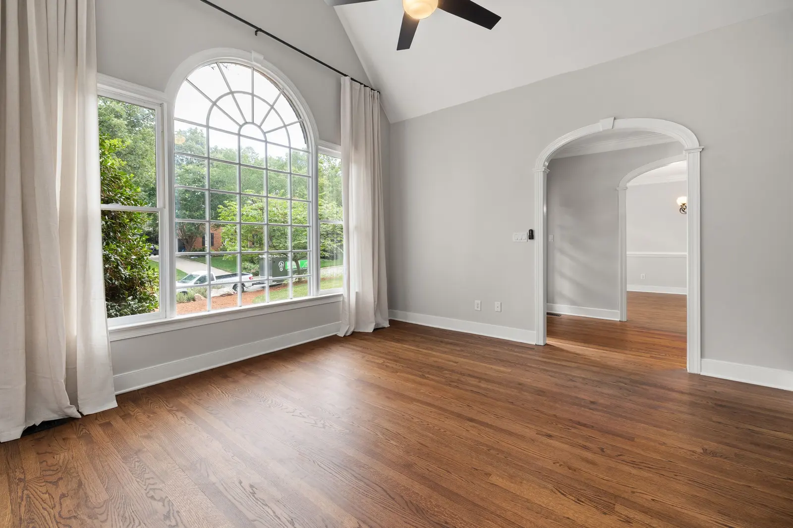 Oak hardwood floor running through an empty living room with a tall arched window and an open archway