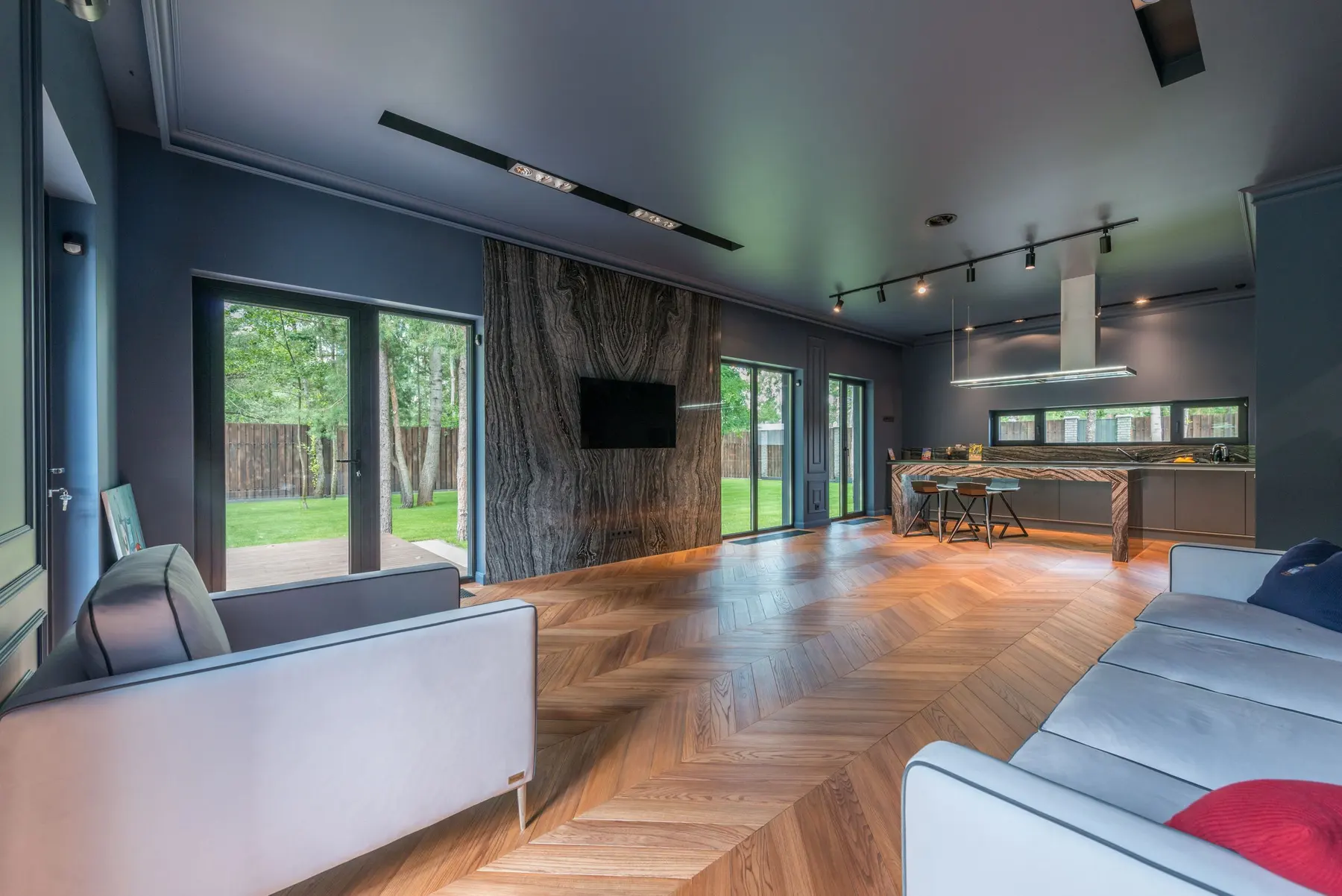 Pale herringbone wood floor in an open-plan living room with a dark ceiling and a kitchen beyond
